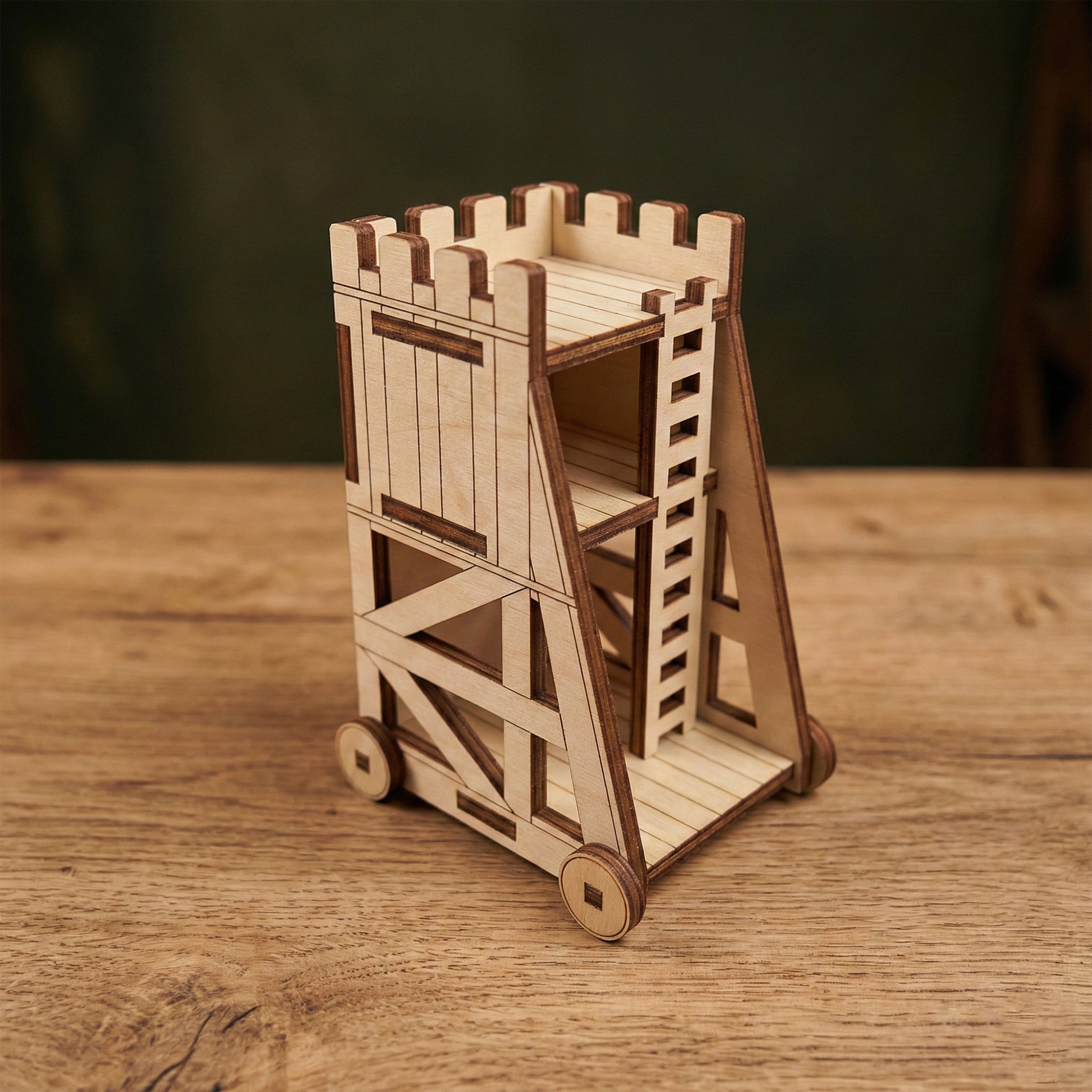 A miniature medieval siege tower made of laser cut plywood. The model features a detailed ladder, crenelated top, and functional-looking wheels. It sits on a wooden surface, showcasing the precise burnt edges and light wood grain.