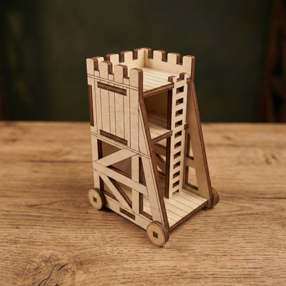 A miniature medieval siege tower made of laser cut plywood. The model features a detailed ladder, crenelated top, and functional-looking wheels. It sits on a wooden surface, showcasing the precise burnt edges and light wood grain.