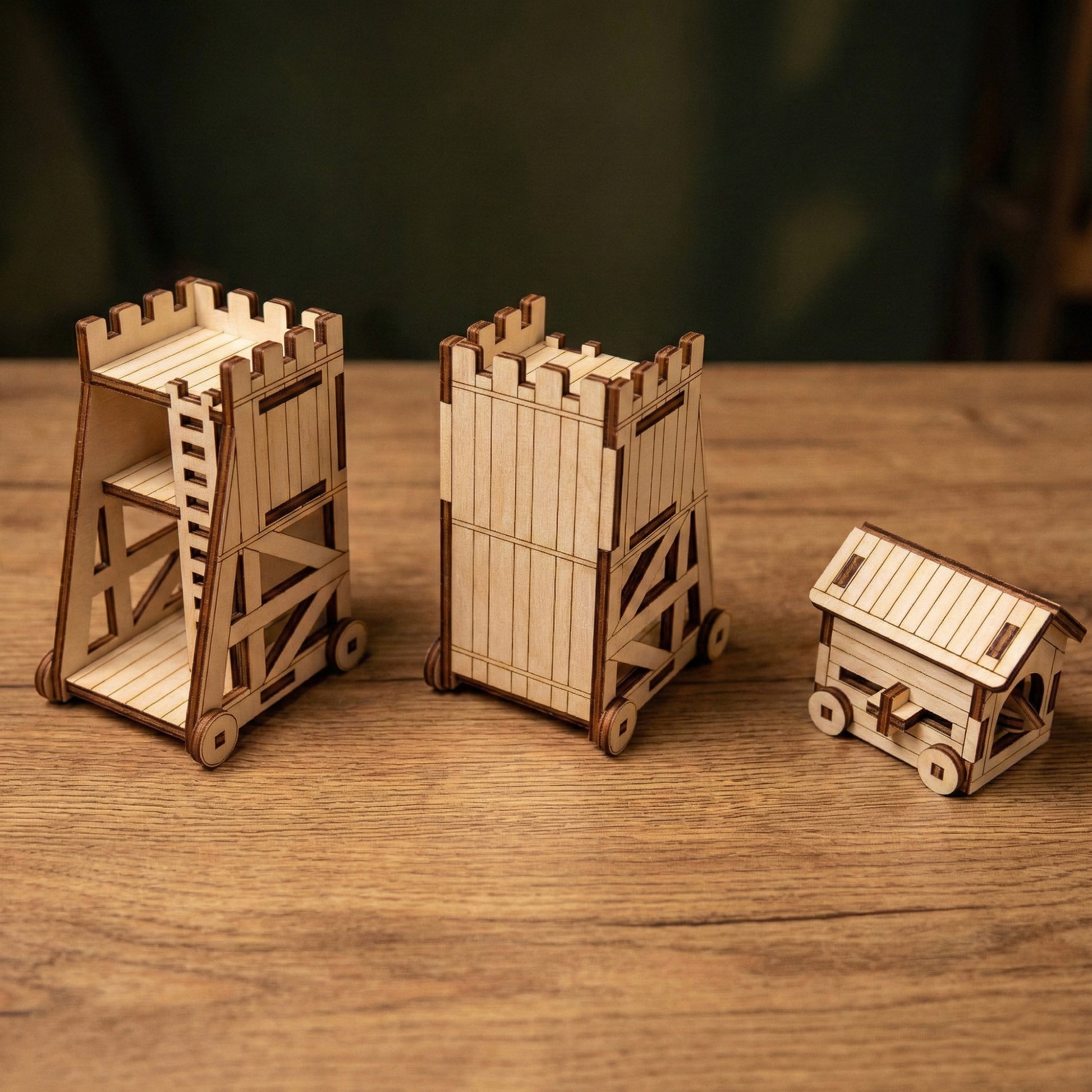 Three different medieval siege weapon models crafted from laser cut plywood. Includes two variations of a tall siege tower and one covered battering ram. The set is displayed in a row on a rustic wooden tabletop.
