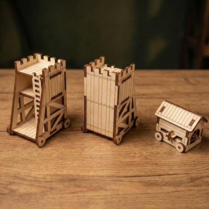 Three different medieval siege weapon models crafted from laser cut plywood. Includes two variations of a tall siege tower and one covered battering ram. The set is displayed in a row on a rustic wooden tabletop.