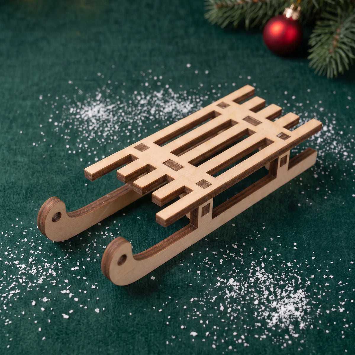 Miniature laser cut plywood sled on green velvet with faux snow flakes, pine needles, and red ornament.