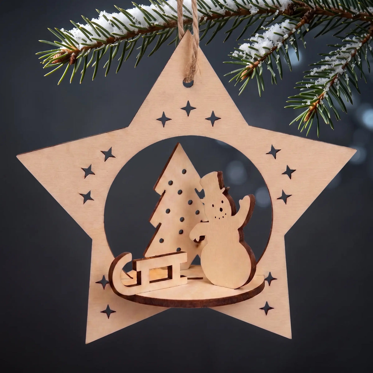 Laser cut plywood star ornament with 3D snowman and sled scene hanging from a snow-dusted pine bough on dark backdrop