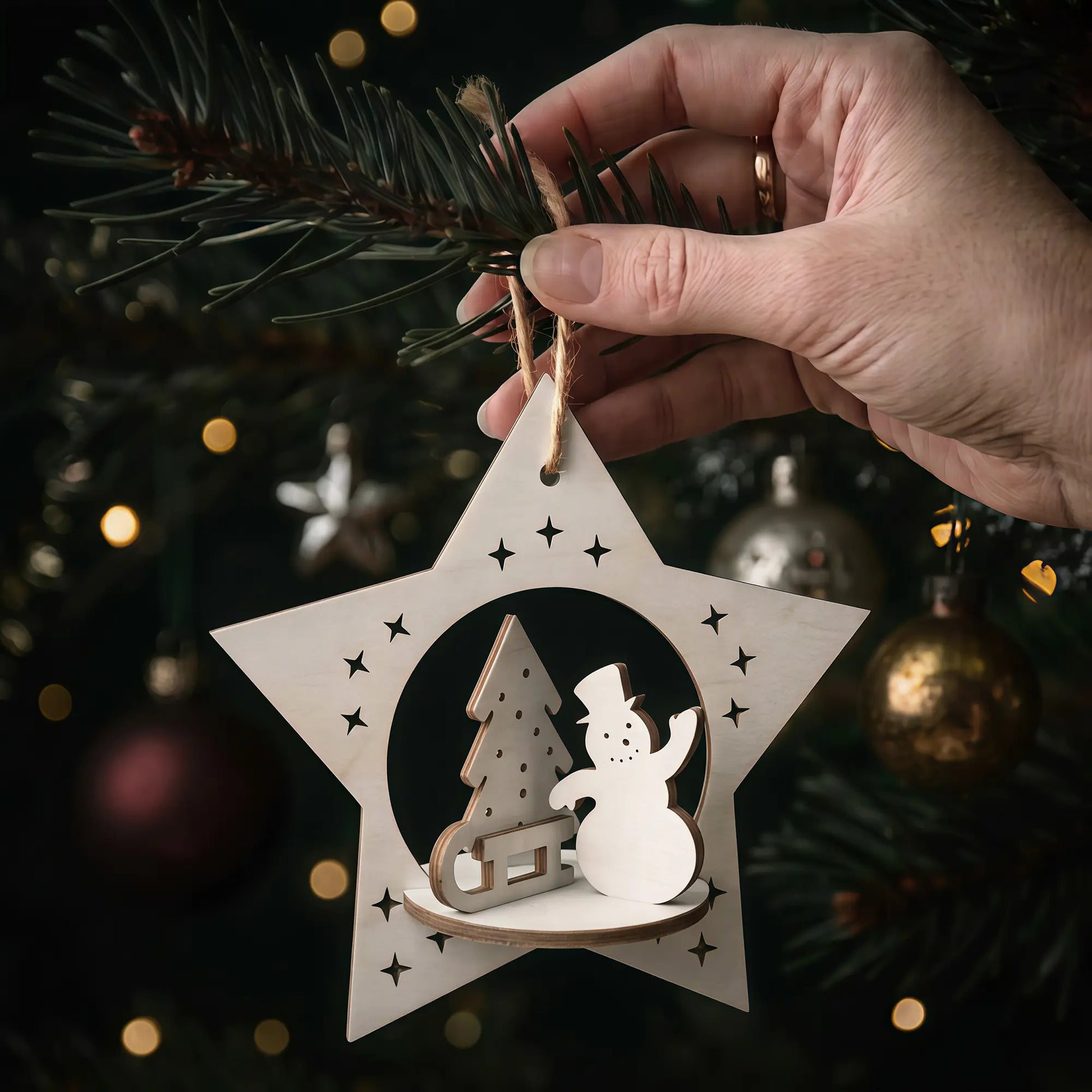 Hand holding laser cut plywood star holiday ornament featuring a 3D painted snowman and sled on a glowing pine tree.