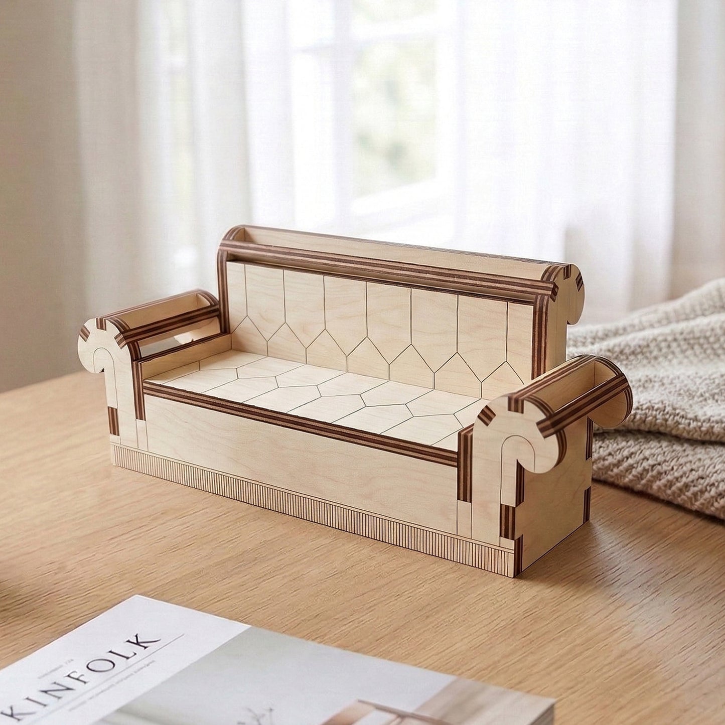 Miniature sofa made of laser cut plywood featuring intricate geometric etchings on the seat and back. Styled with elegant scroll-shaped armrests and a striped base detail. Displayed on a wooden surface next to a magazine