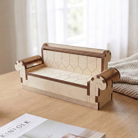 Miniature sofa made of laser cut plywood featuring intricate geometric etchings on the seat and back. Styled with elegant scroll-shaped armrests and a striped base detail. Displayed on a wooden surface next to a magazine