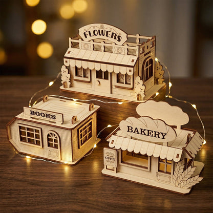 A trio of miniature storefront ornaments—Flowers, Books, and Bakery—crafted from laser-cut plywood. The natural wood models feature layered details, tiny awnings, and engraved signage. Displayed on wooden blocks with warm fairy lights for a cozy aesthetic.