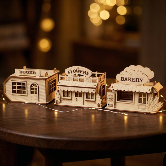 A row of three miniature wooden shops (Books, Flowers, and Bakery) made from light-colored plywood with dark burnt edges. Each model highlights intricate window frames and decorative storefront trim. Posed on a dark wood table with glowing ambient lights.