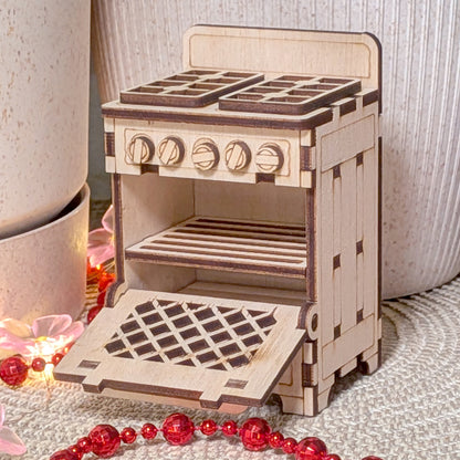 Laser cut wooden miniature dollhouse stove with opening oven door, detailed knobs and burners, displayed indoors with decorative beads and soft lighting.