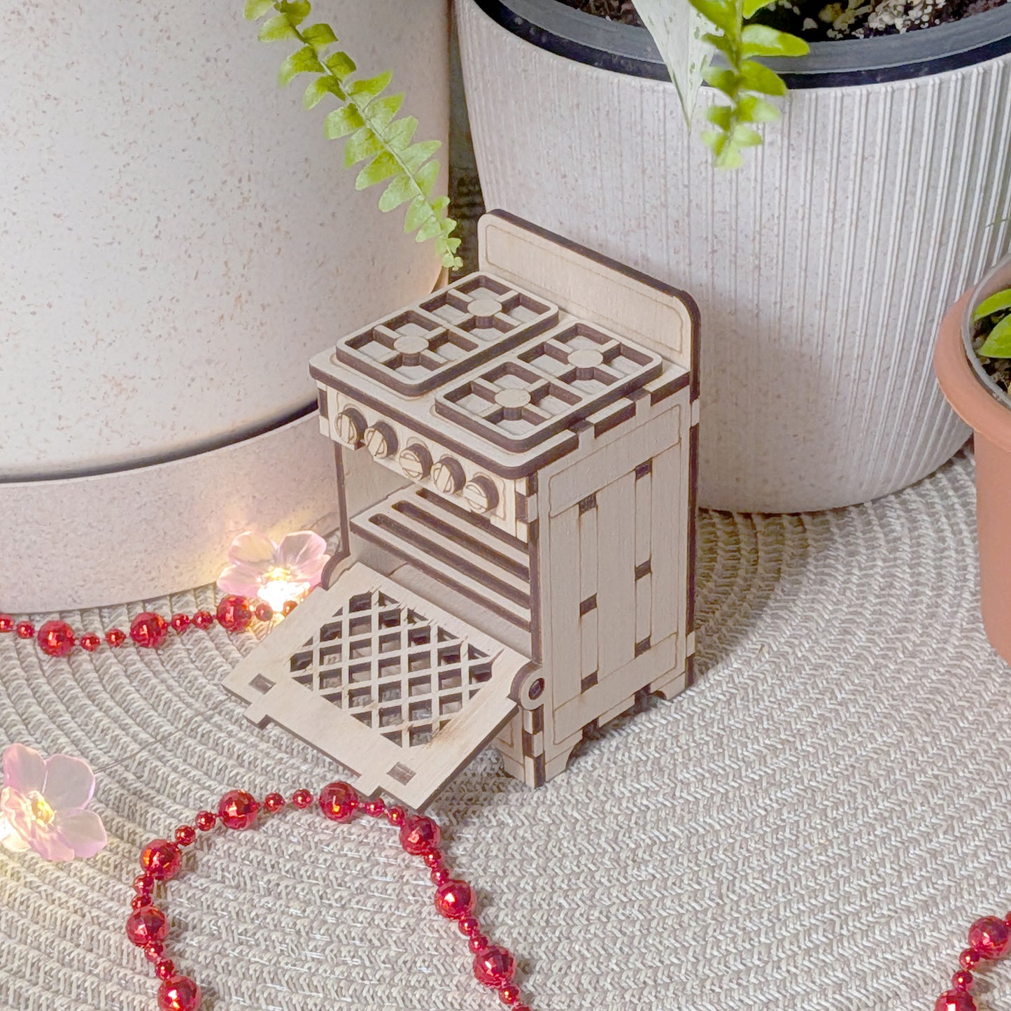 Laser cut wooden miniature stove with the oven door open, displayed on a textured surface with decorative lights and plants.