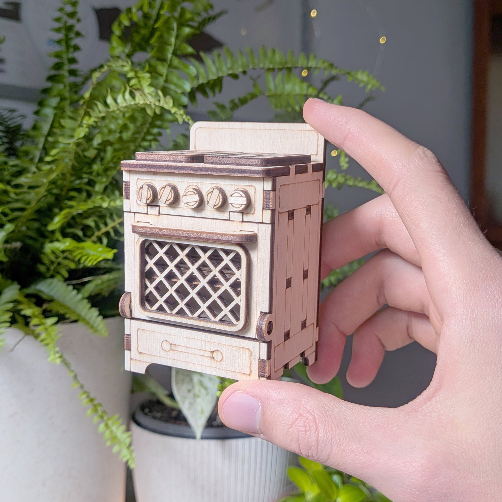 Laser cut wooden miniature stove held at an angle, showing engraved knobs, lattice oven door, and panel details