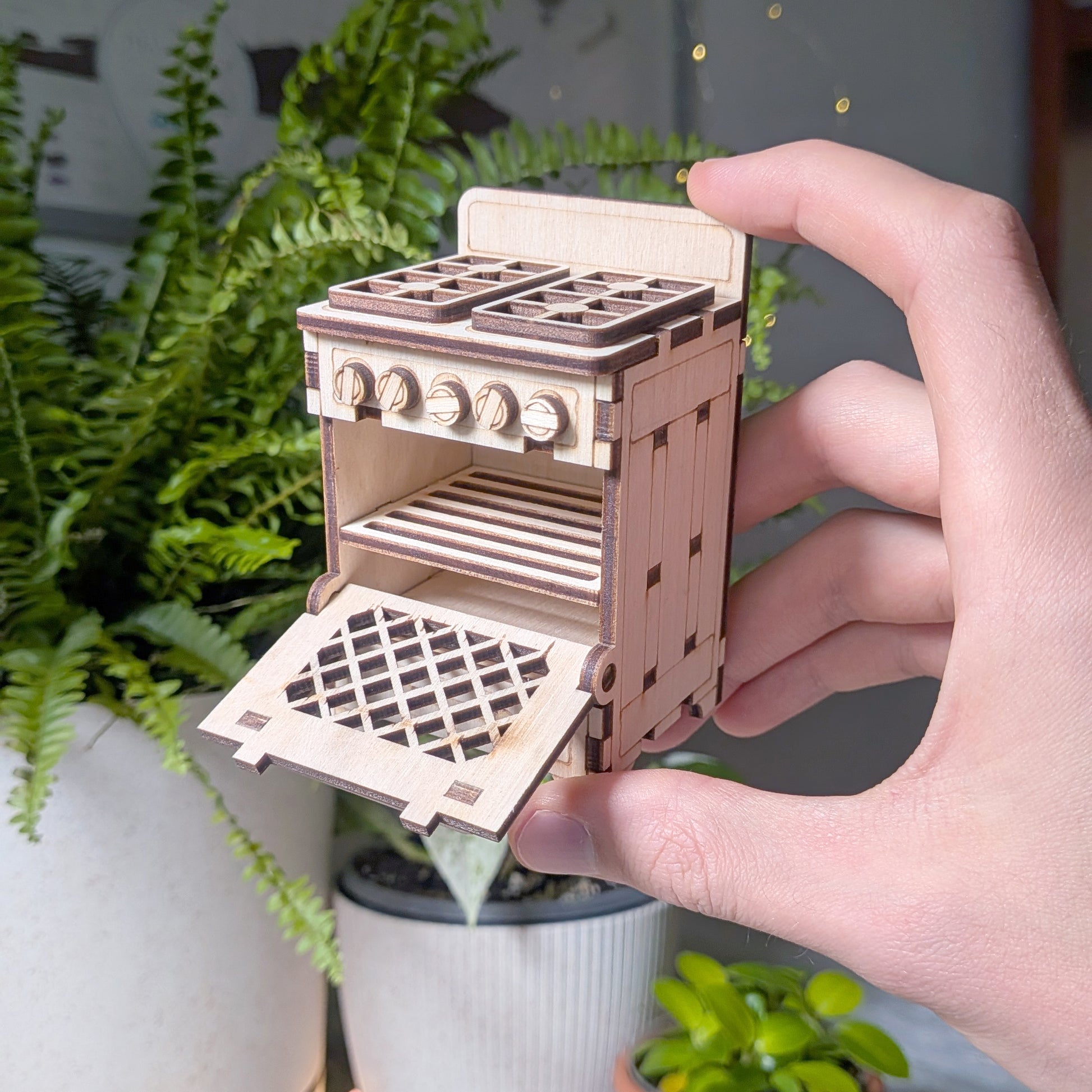 Laser cut wooden dollhouse stove held in a hand with the oven door open, showing the interior shelf and detailed plywood construction.