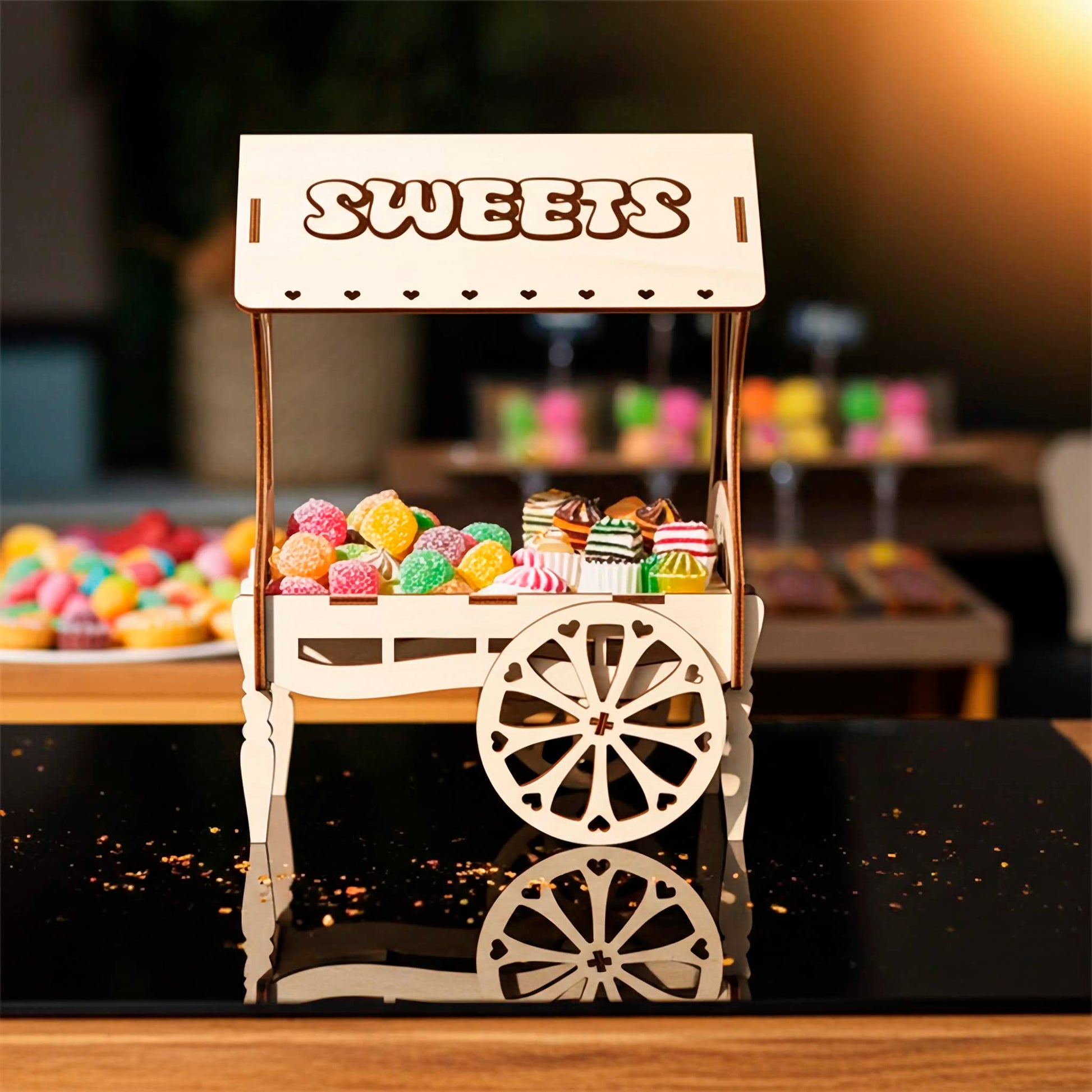 A light wooden plywood laser cut miniature vendor cart labeled "SWEETS" on the roof, filled with colorful jelly candies, resting on a reflective black surface with glitter, taken in bright outdoor lighting.