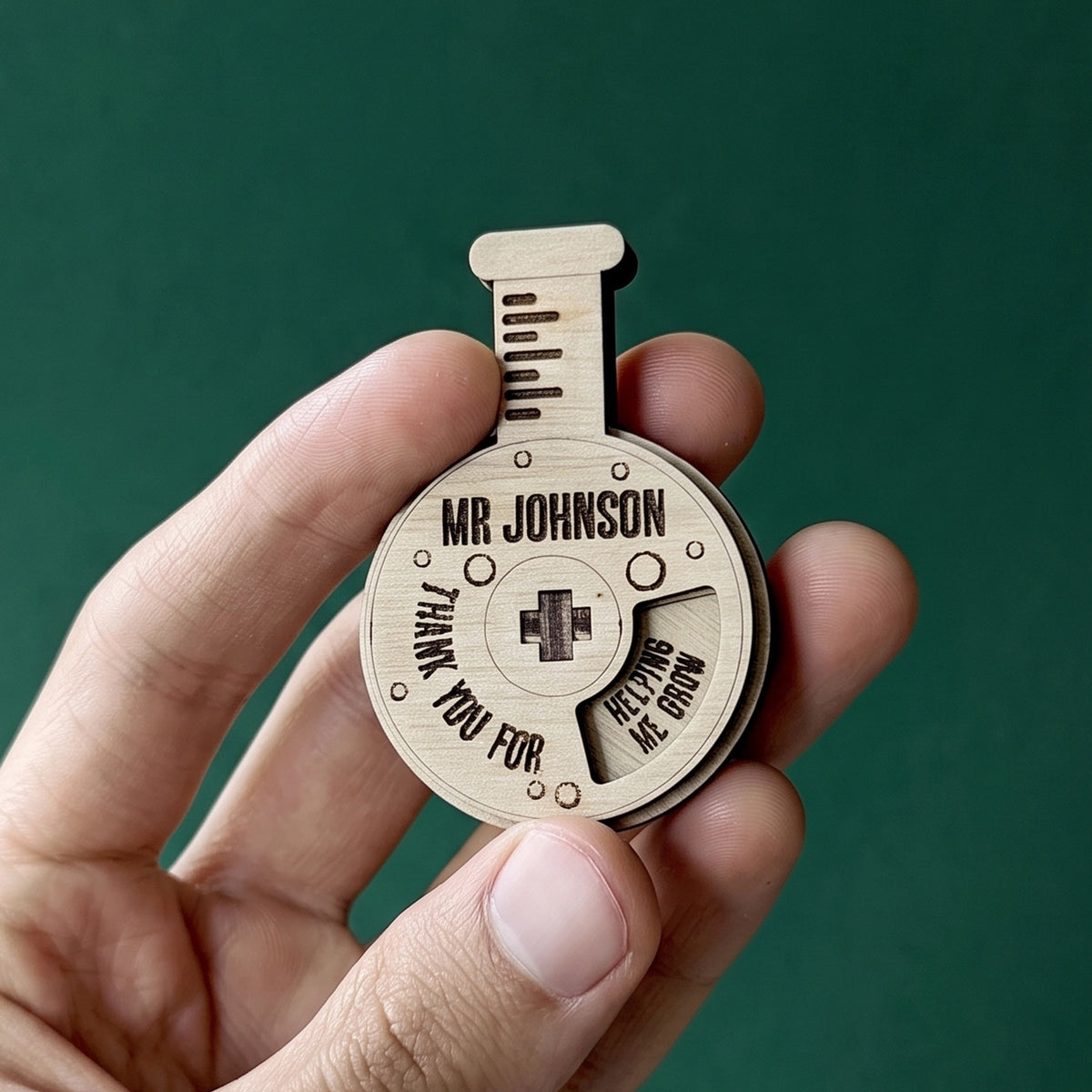 Hand holding a personalized laser cut plywood chemistry flask teacher appreciation spinner against dark green background.