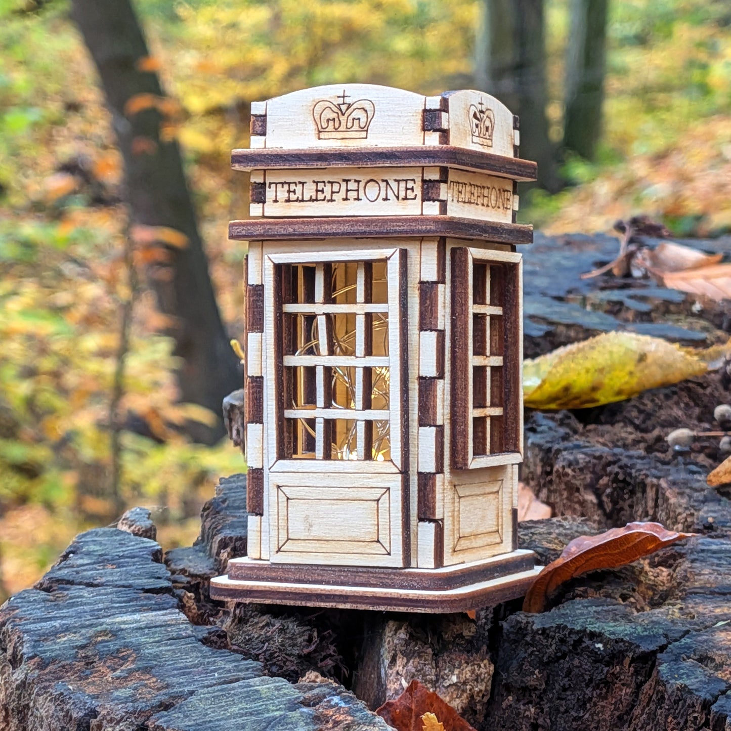Laser cut wooden telephone booth miniature displayed outdoors on a tree stump, featuring engraved crown symbols, window grids, and layered plywood construction.