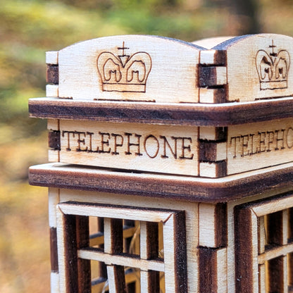 Close-up of a laser cut wooden miniature telephone booth showing engraved crown details and the word “TELEPHONE” on the top panel, with light and dark plywood layers.