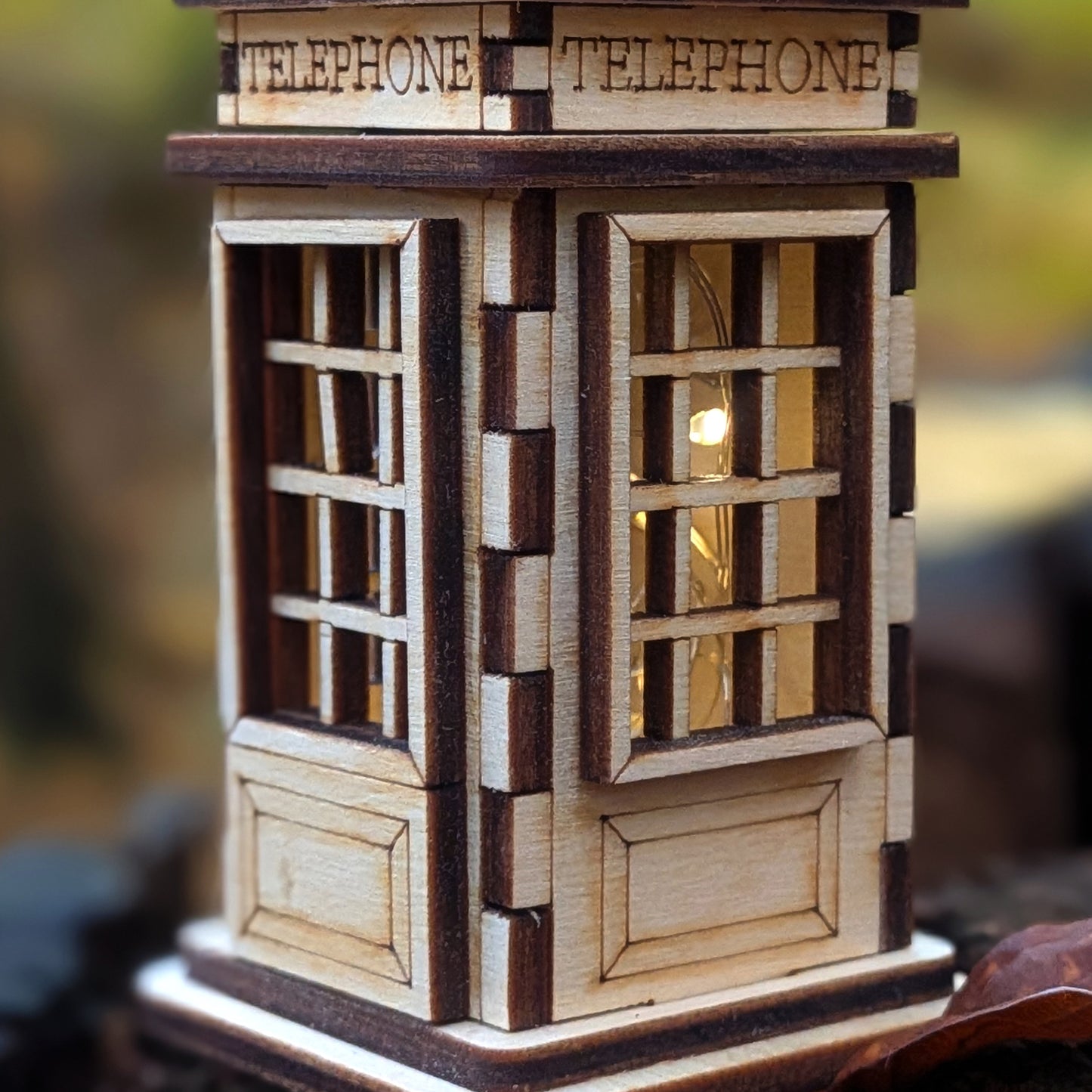Laser cut wooden telephone booth miniature with detailed window grids and warm light glowing inside, crafted from layered light and dark plywood.