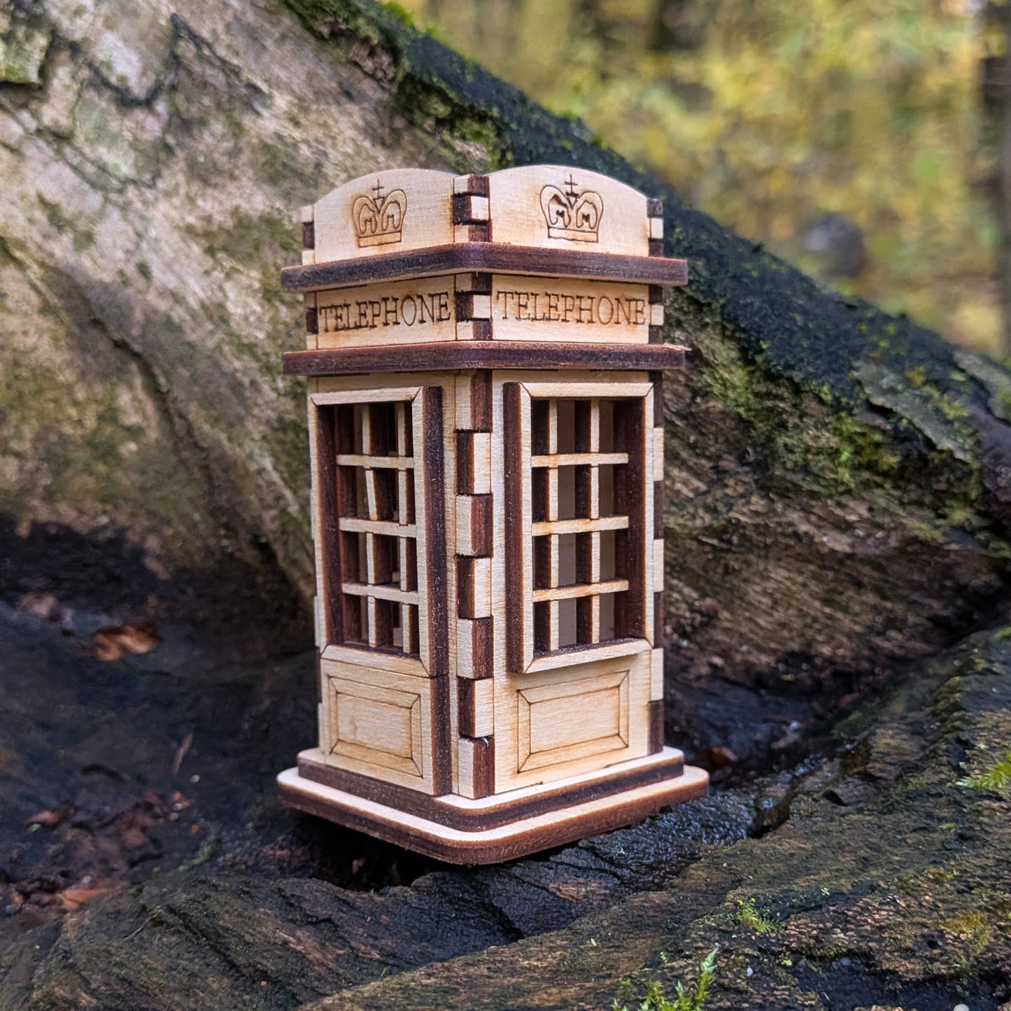 Laser cut wooden telephone booth miniature resting on mossy wood, showing detailed engraved panels and window grids in light and dark plywood.
