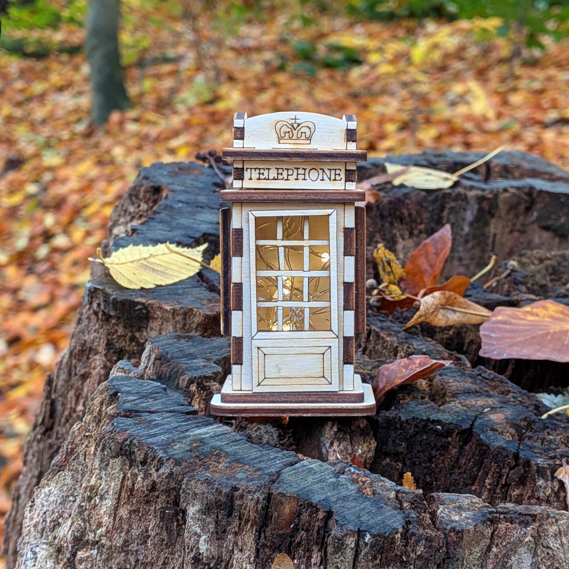 Front view of a laser cut wooden telephone booth miniature standing on a tree stump surrounded by autumn leaves, with engraved panels and window frames.