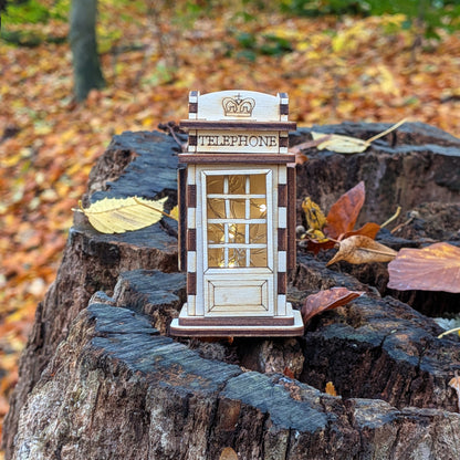 Front view of a laser cut wooden telephone booth miniature standing on a tree stump surrounded by autumn leaves, with engraved panels and window frames.