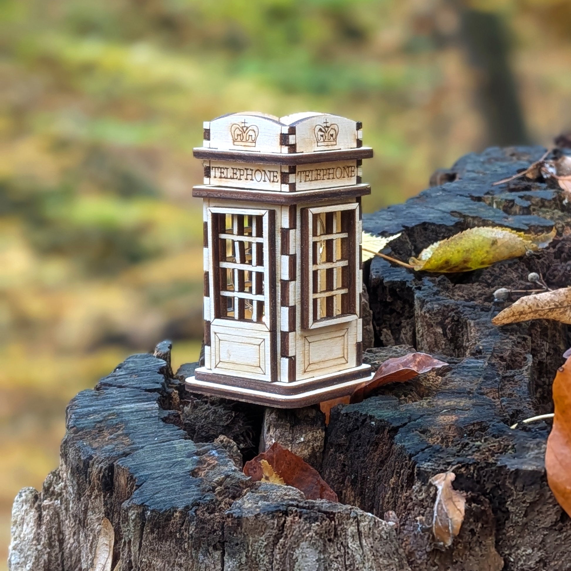 Laser cut wooden miniature telephone booth displayed outdoors on a cracked tree stump, showing engraved details and layered plywood construction.