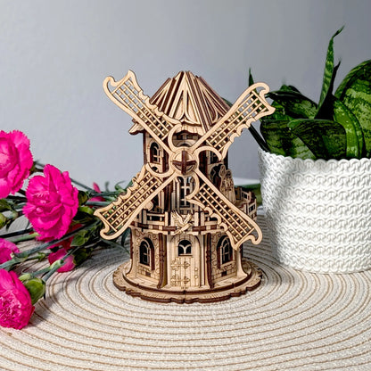 Straight front view of a laser cut plywood windmill model on a woven mat next to bright pink flowers and a houseplant.