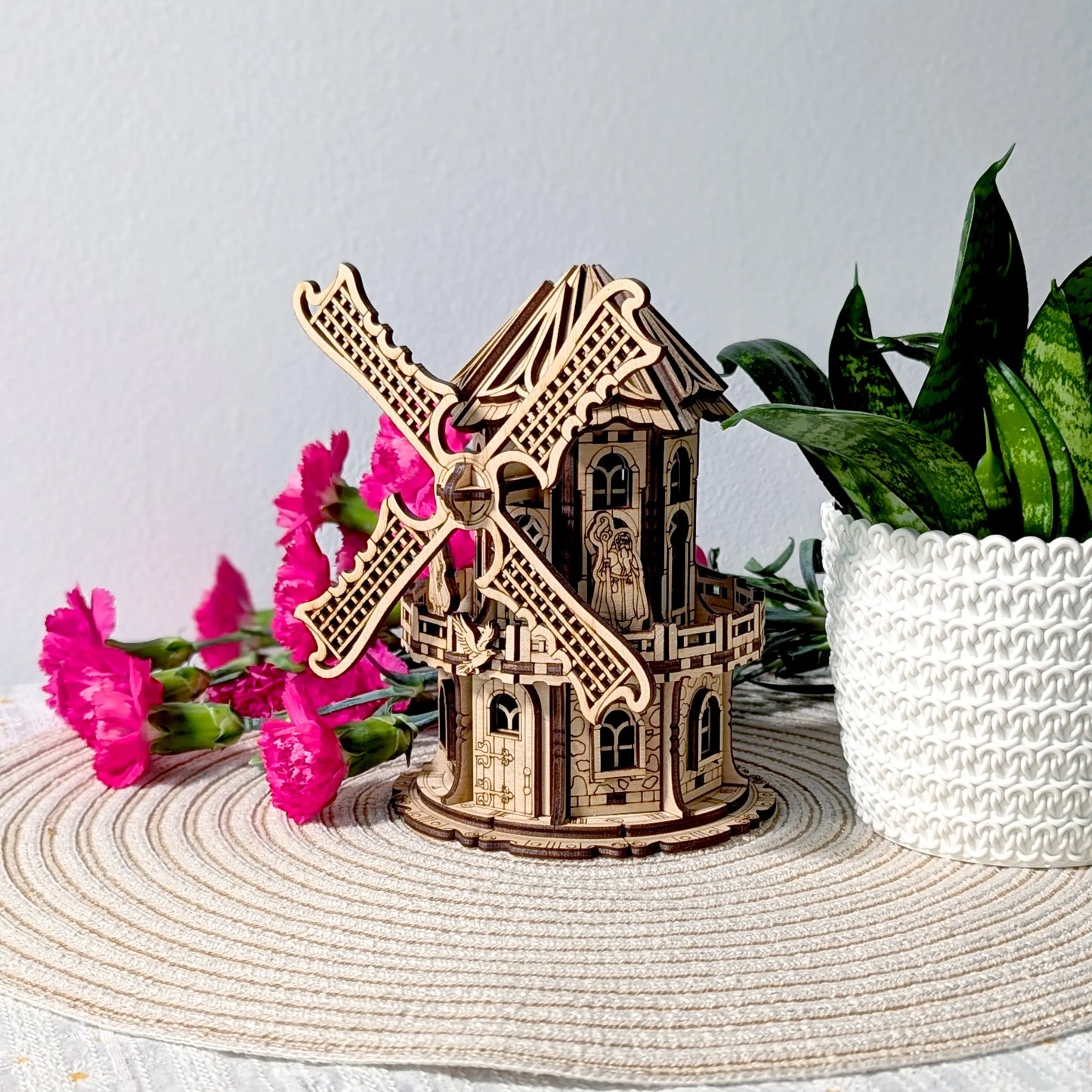 Angled profile view of a finished laser cut plywood windmill model staged nicely with bright pink flowers and a plant.