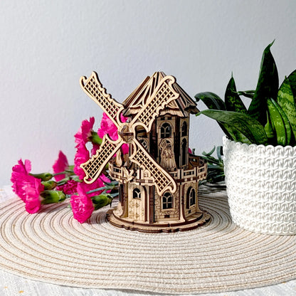 Angled profile view of a finished laser cut plywood windmill model staged nicely with bright pink flowers and a plant.