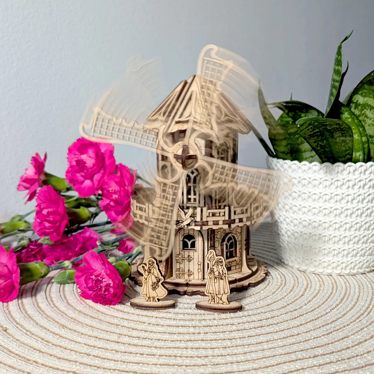 Wooden laser cut model of a windmill with two figures and pink flowers on a textured surface.