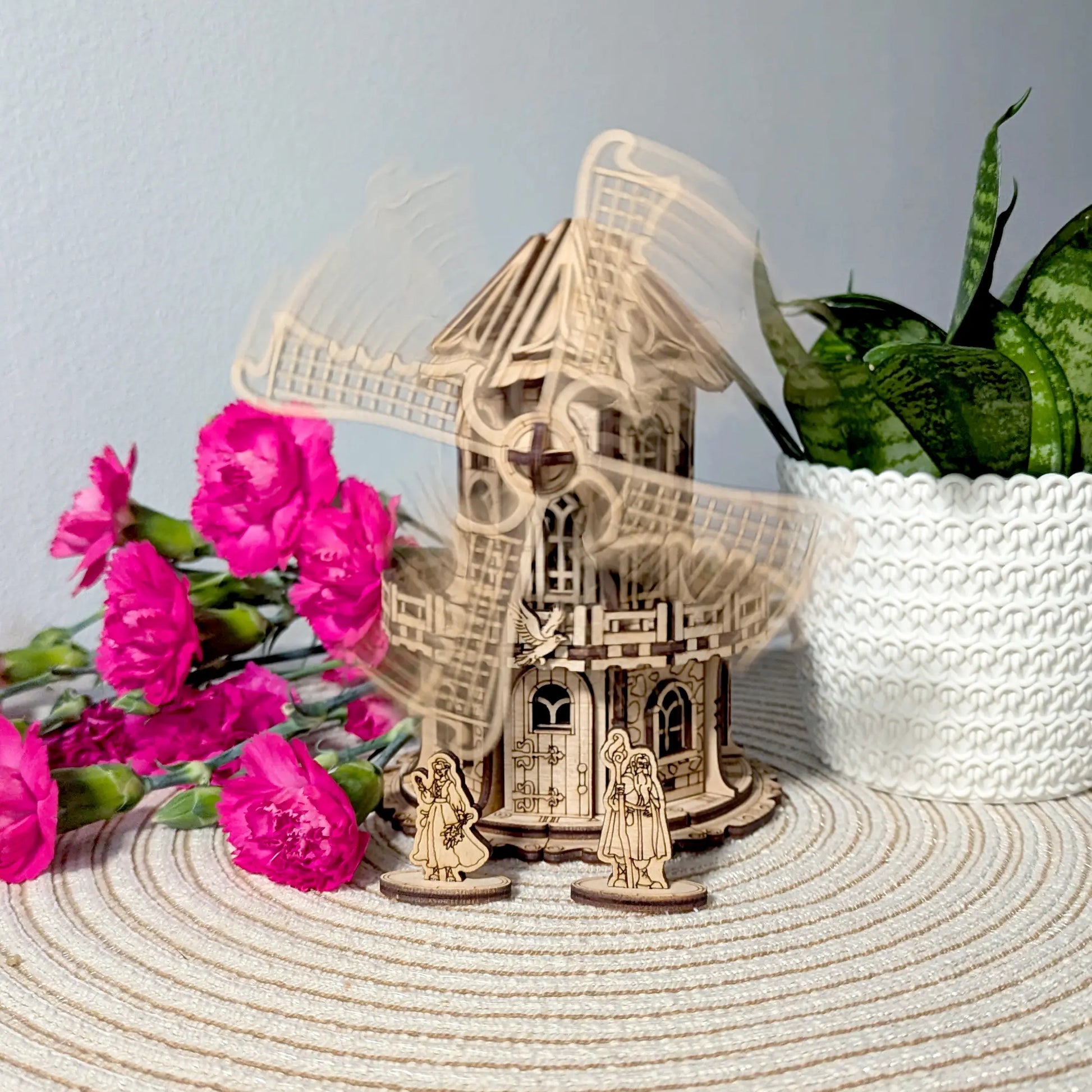 Wooden laser cut model of a windmill with two figures and pink flowers on a textured surface.