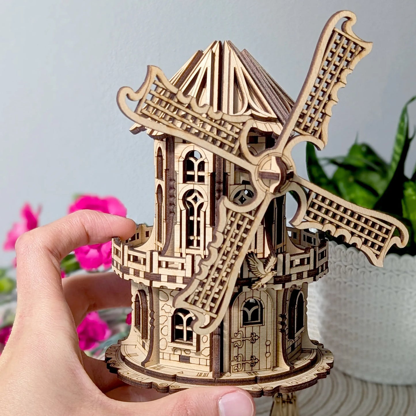 A hand holding a laser-cut wooden model of a windmill, with intricate details and curved blades.