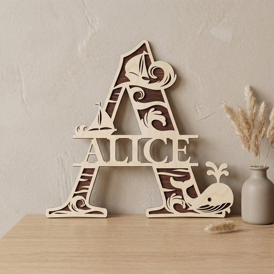 A personalized laser-cut wooden monogram letter A featuring the name "Alice." The two-toned plywood design has a nautical theme with ocean waves, sailboats, and a spouting whale. Displayed on a light wood table against a textured wall with a vase of dried pampas grass.