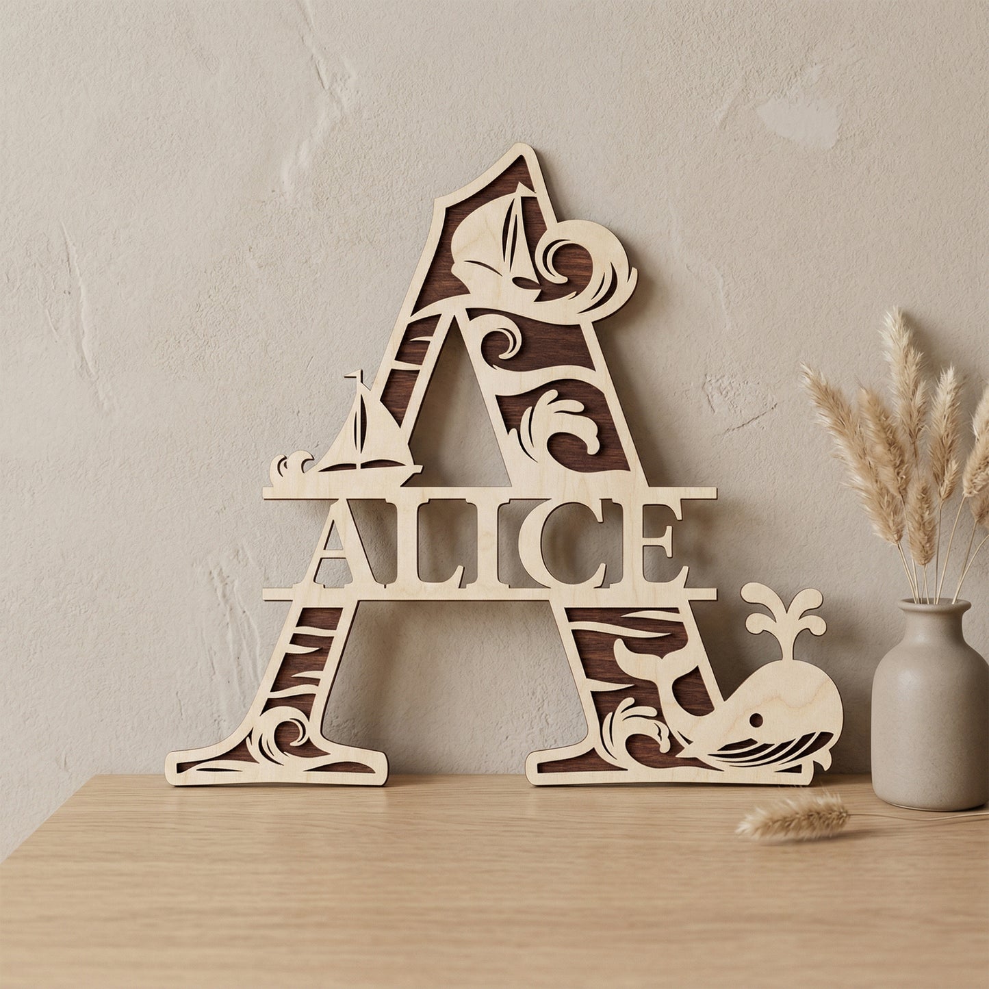 A personalized laser-cut wooden monogram letter A featuring the name "Alice." The two-toned plywood design has a nautical theme with ocean waves, sailboats, and a spouting whale. Displayed on a light wood table against a textured wall with a vase of dried pampas grass.