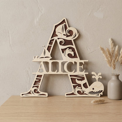 A personalized laser-cut wooden monogram letter A featuring the name "Alice." The two-toned plywood design has a nautical theme with ocean waves, sailboats, and a spouting whale. Displayed on a light wood table against a textured wall with a vase of dried pampas grass.