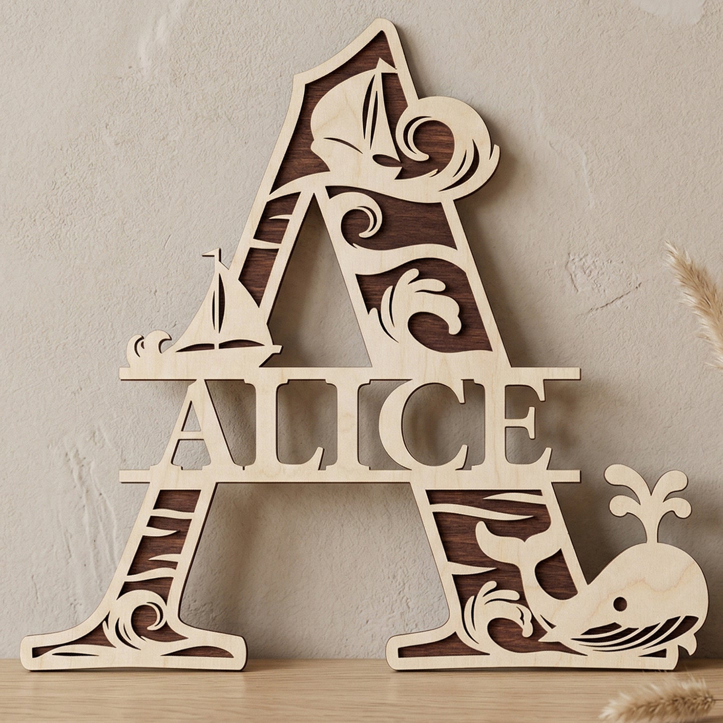 A personalized laser-cut wooden monogram letter A featuring the name "Alice." The two-toned plywood design has a nautical theme with ocean waves, sailboats, and a spouting whale. Displayed on a light wood table against a textured wall with a vase of dried pampas grass.