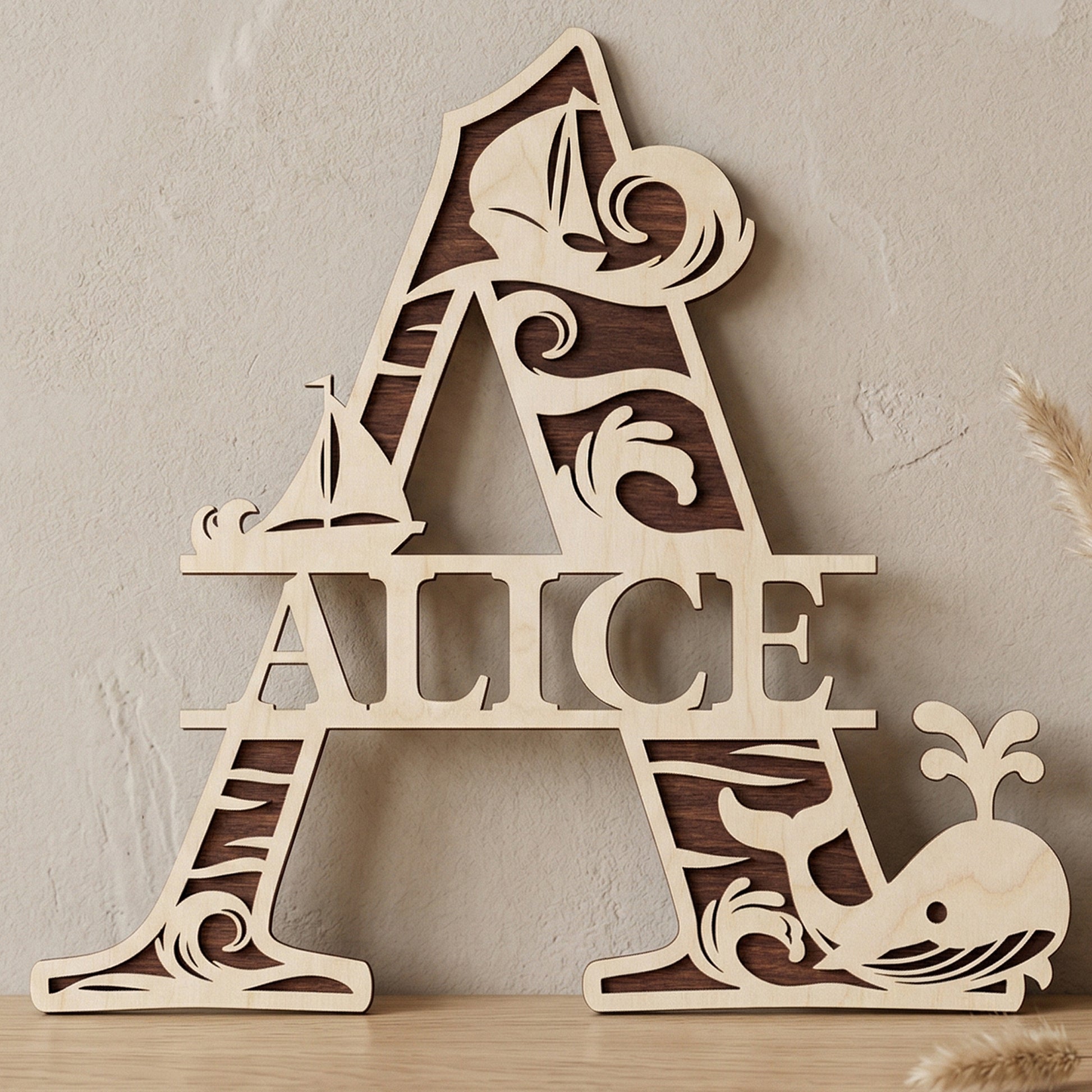 A personalized laser-cut wooden monogram letter A featuring the name "Alice." The two-toned plywood design has a nautical theme with ocean waves, sailboats, and a spouting whale. Displayed on a light wood table against a textured wall with a vase of dried pampas grass.