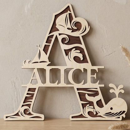 A personalized laser-cut wooden monogram letter A featuring the name "Alice." The two-toned plywood design has a nautical theme with ocean waves, sailboats, and a spouting whale. Displayed on a light wood table against a textured wall with a vase of dried pampas grass.