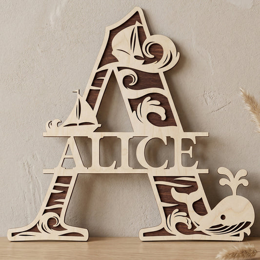 A personalized laser-cut wooden monogram letter A featuring the name "Alice." The two-toned plywood design has a nautical theme with ocean waves, sailboats, and a spouting whale. Displayed on a light wood table against a textured wall with a vase of dried pampas grass.