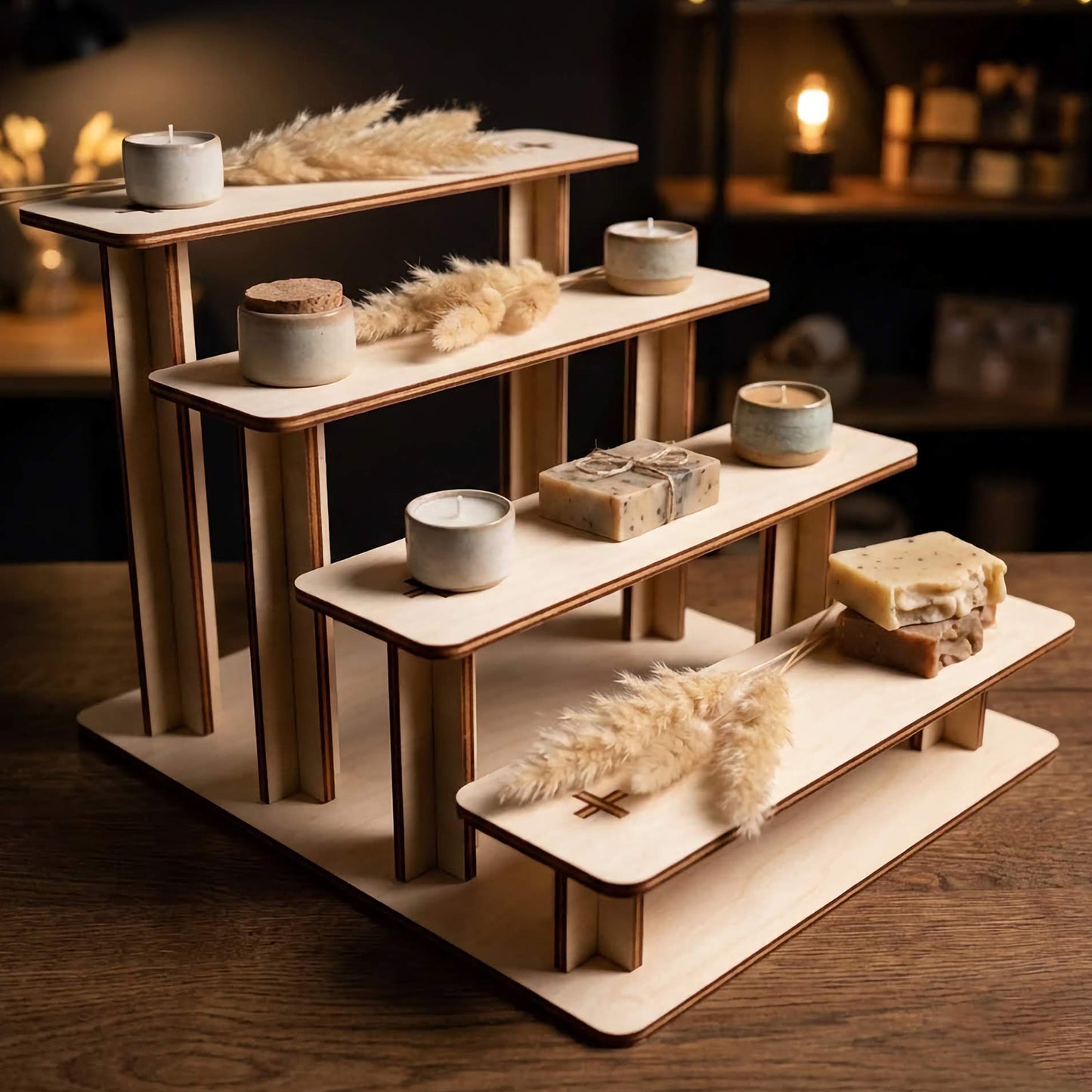 Four-tiered laser cut plywood riser in a dimly lit room, holding ceramic candles, handmade soaps, and dried pampas grass. Warm ambient lighting highlights the smooth wood texture and tiered design, perfect for a cozy home or boutique display.