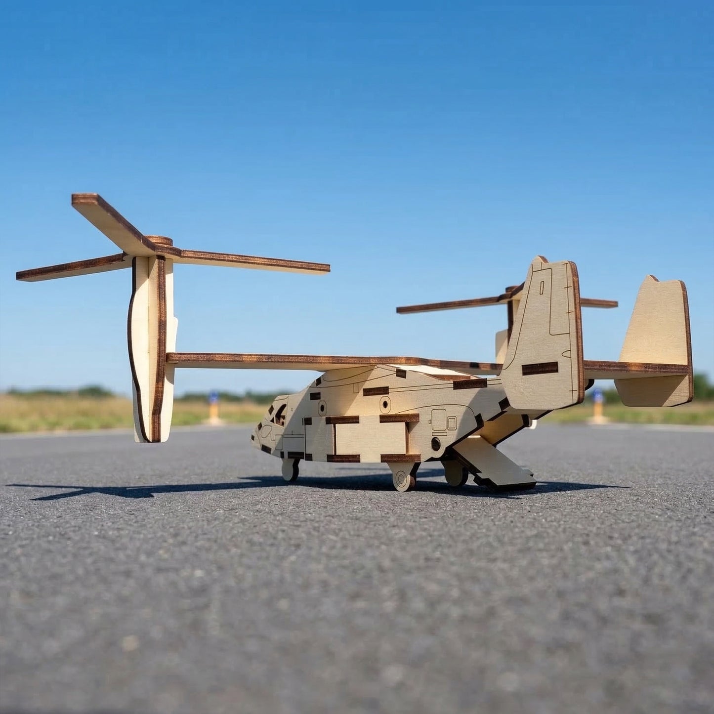 Rear-view of a laser cut plywood tiltrotor aircraft model with its back cargo ramp open. Detailed wooden assembly showing interlocking slots, twin vertical stabilizers, and long wings resting on a sunny airport tarmac under a blue sky.