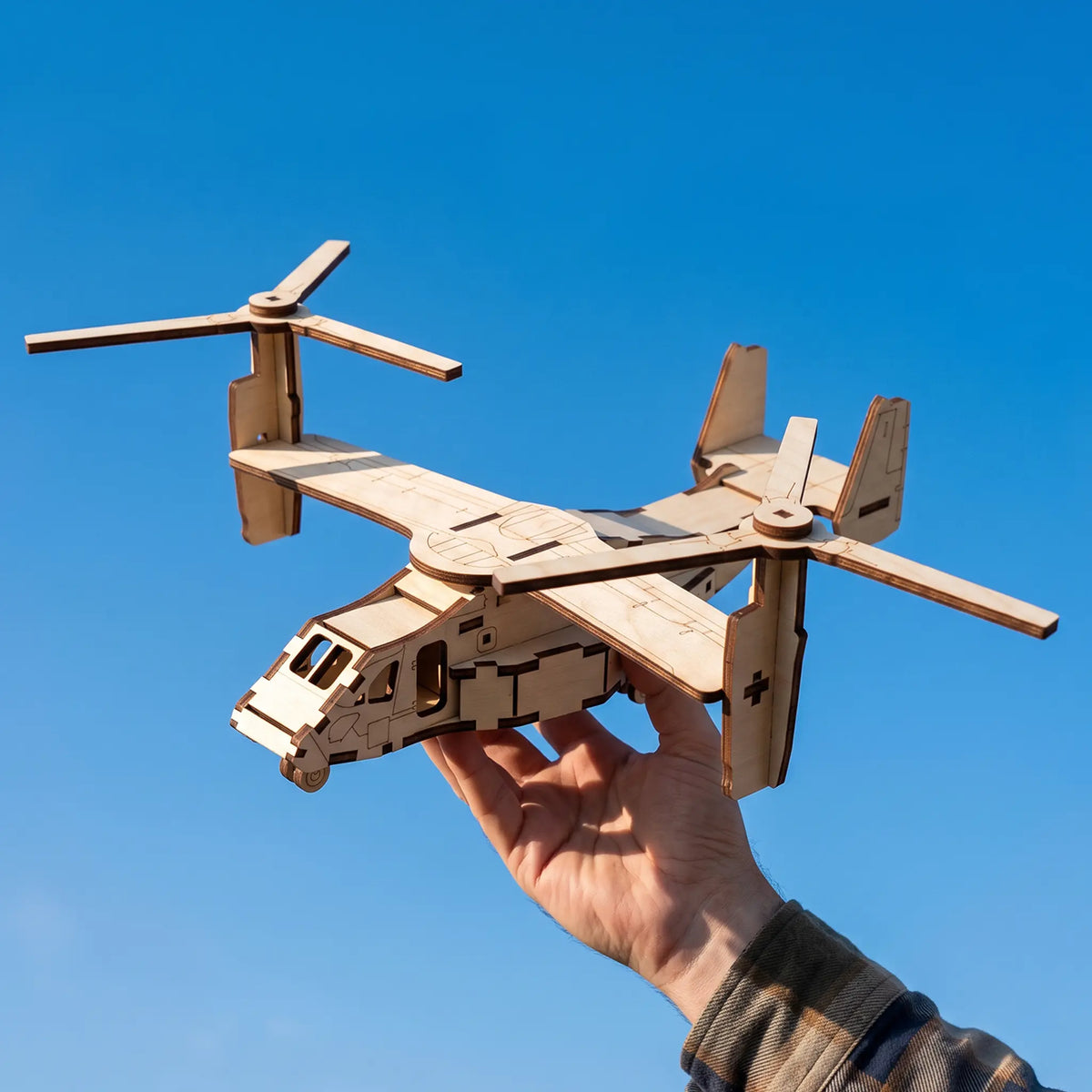 Hand holding assembled laser cut plywood tiltrotor aircraft model up in the air against a clear blue sunny sky background.
