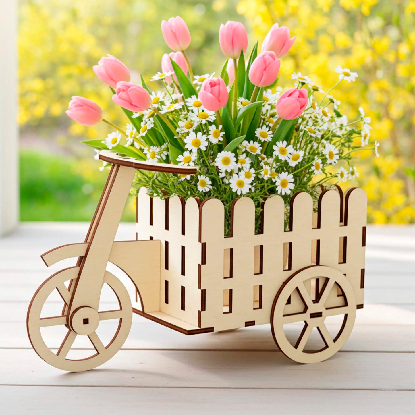 Light wooden plywood laser cut tricycle flower stand with a picket fence style cargo box, filled with pink tulips and daisies. Displayed on a white wooden deck with a bright, blurred garden background, highlighting its intricate wood detail.