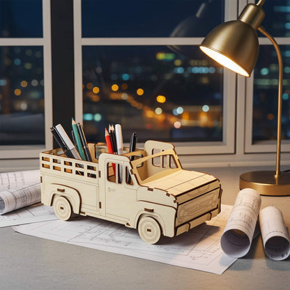 Laser cut wooden plywood pickup truck pencil holder on a desk with blueprints and rolled architectural drawings. The 3D plywood model features a slatted truck bed holding pens and pencils, with a city skyline and desk lamp in the background.