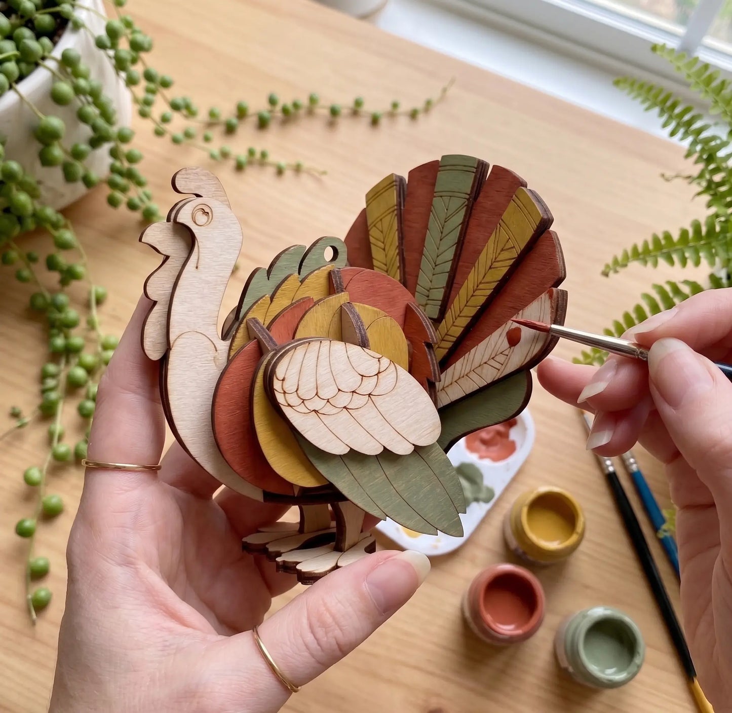 Hand holding a wooden laser cut turkey decoration with paint cans and brushes on a table.