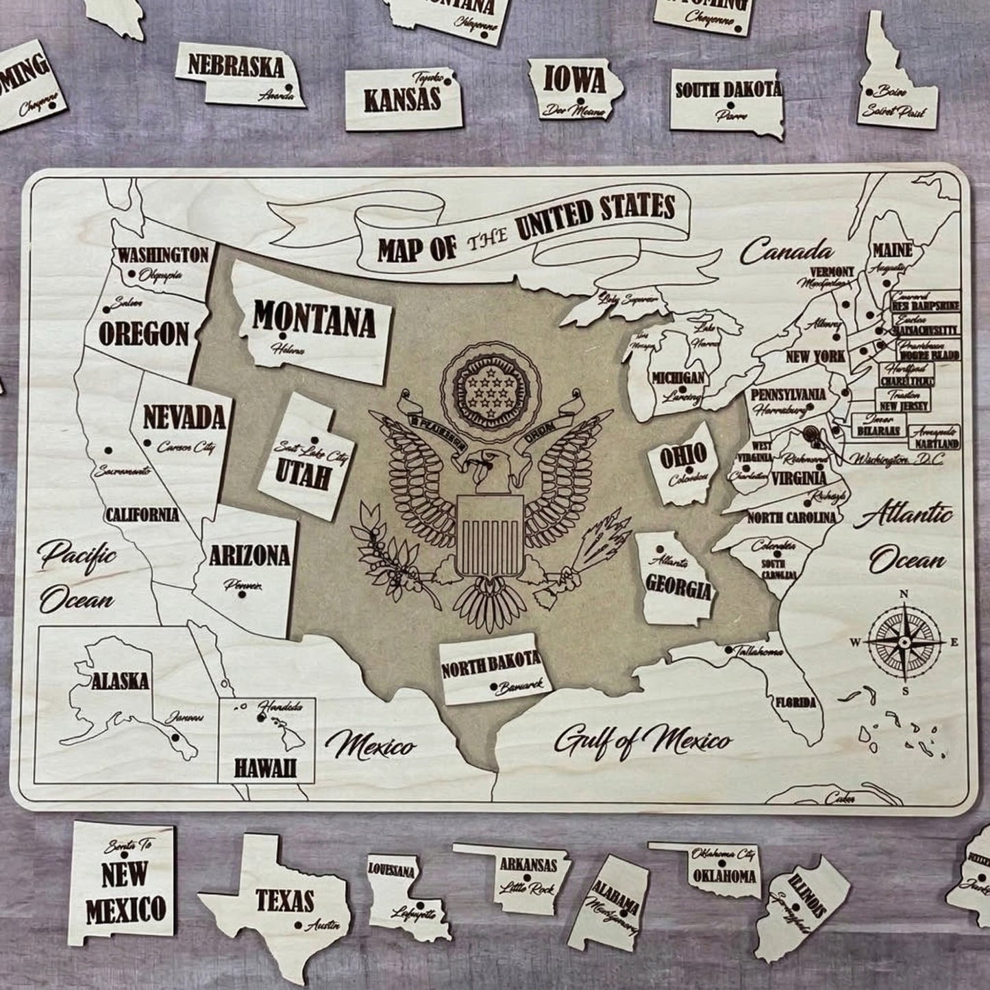 A partially assembled USA puzzle map made of laser cut plywood. Several state pieces like Texas and New Mexico are scattered around a wooden base board that features an engraved eagle crest in the center and a decorative title banner.