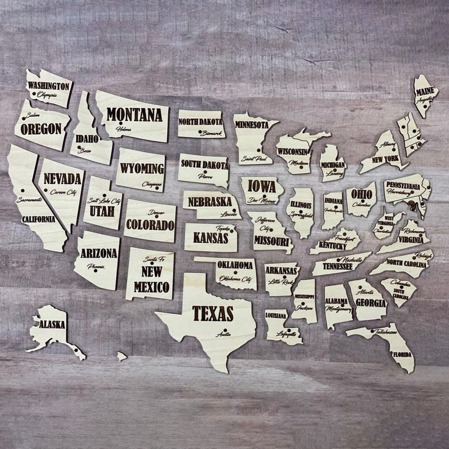 Individual state pieces of a USA map made from laser cut plywood, arranged in geographic order without the base board. Each light wood piece features high-contrast black engraved text for the state name and capital city.