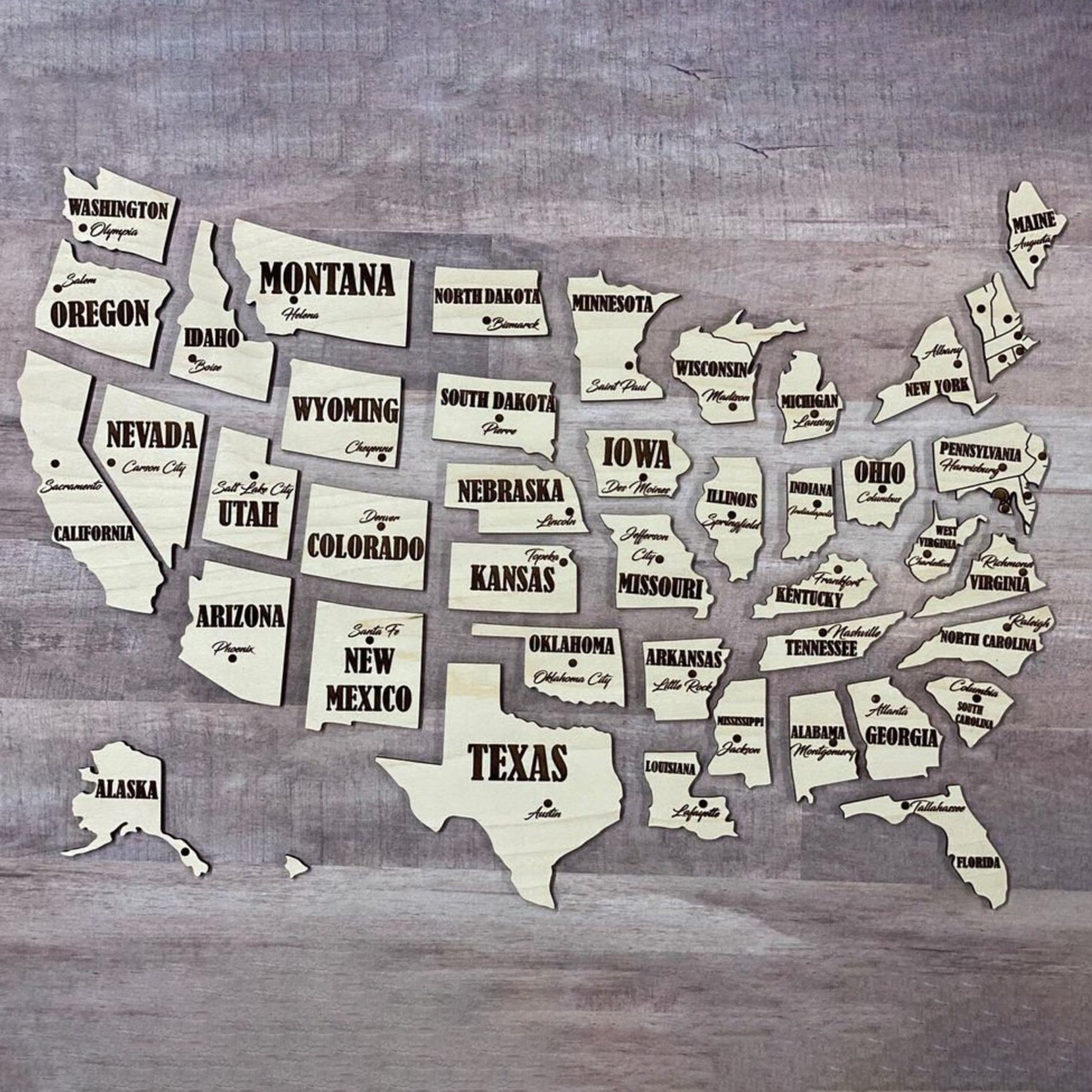 Individual state pieces of a USA map made from laser cut plywood, arranged in geographic order without the base board. Each light wood piece features high-contrast black engraved text for the state name and capital city.