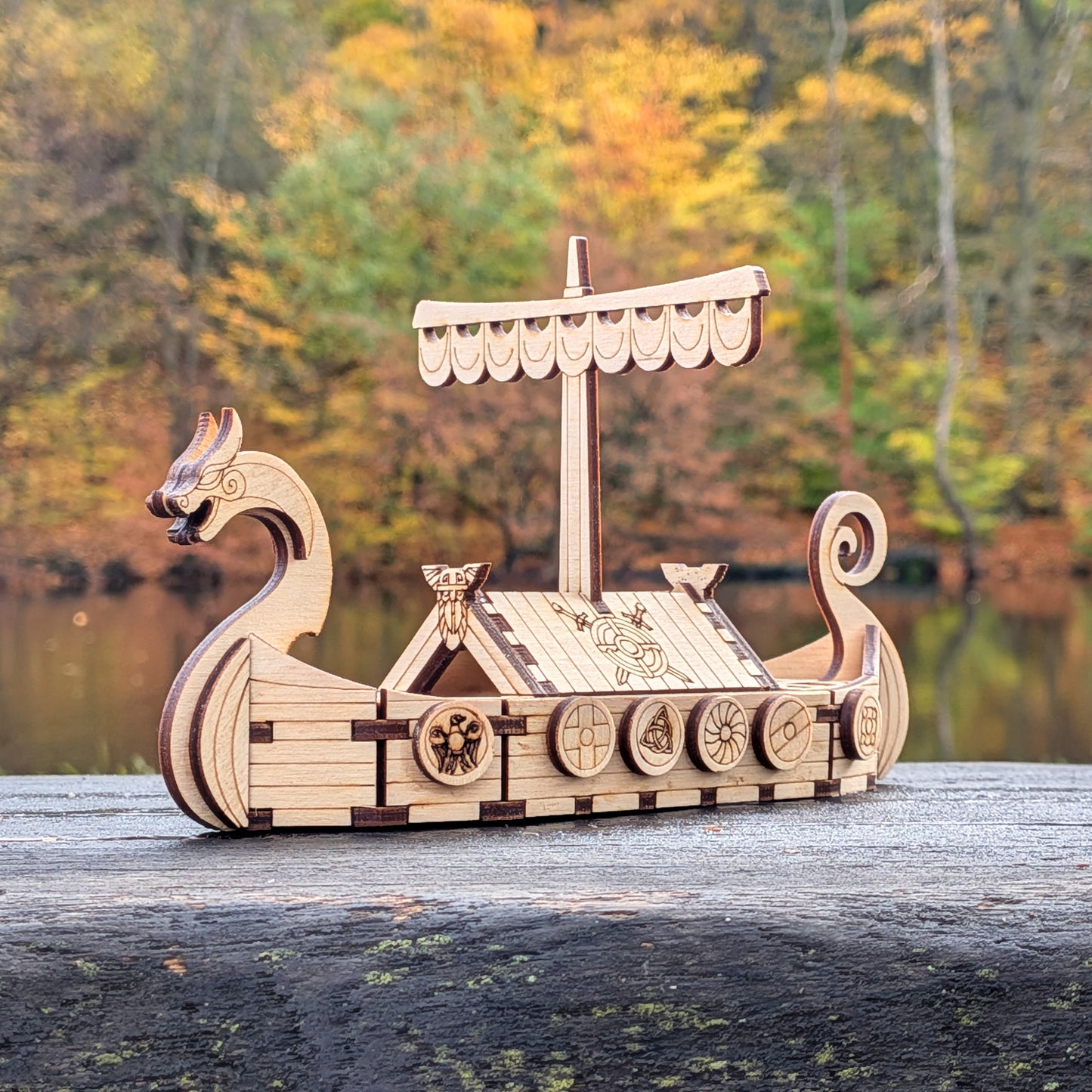 Laser cut wooden Viking ship model displayed outdoors on a wooden surface, with autumn trees and a calm lake in the background.