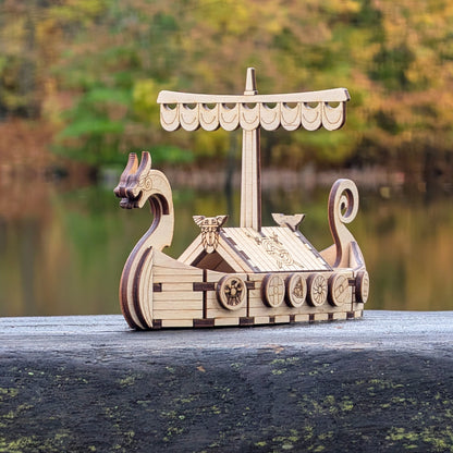 Laser cut wooden Viking ship model displayed outdoors on a wooden surface, with autumn trees and a calm lake in the background