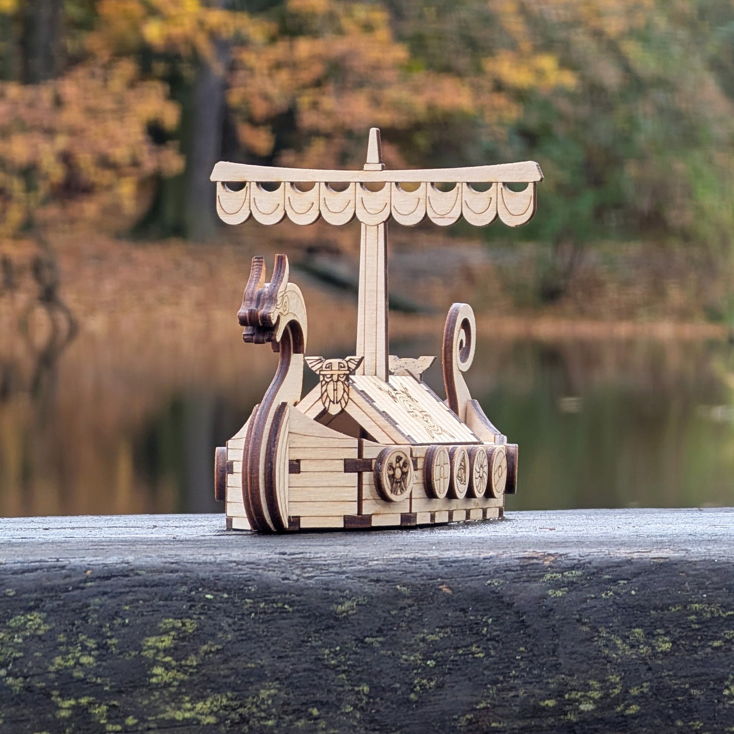 Laser cut wood Viking longship model resting on a weathered wooden board outdoors, showing curved prow and patterned shields.