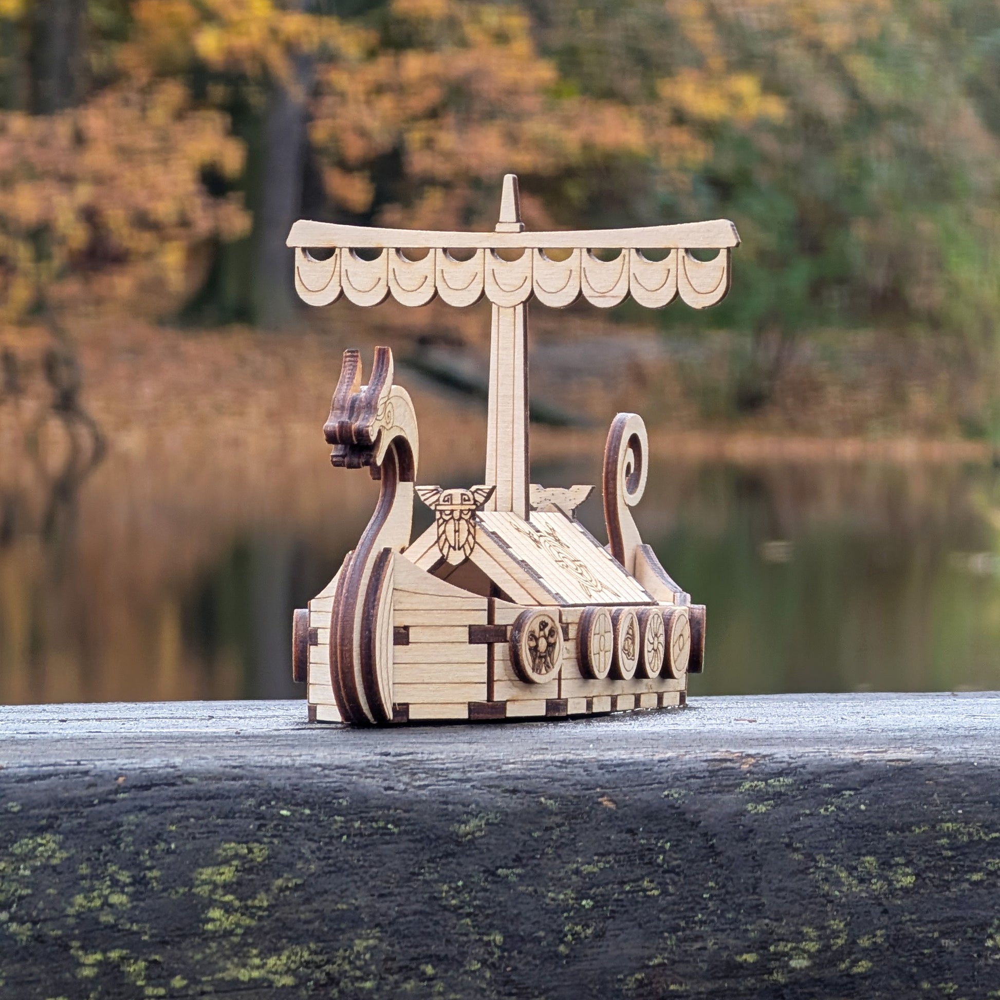 Laser cut wood Viking longship model resting on a weathered wooden board outdoors, showing curved prow and patterned shields.